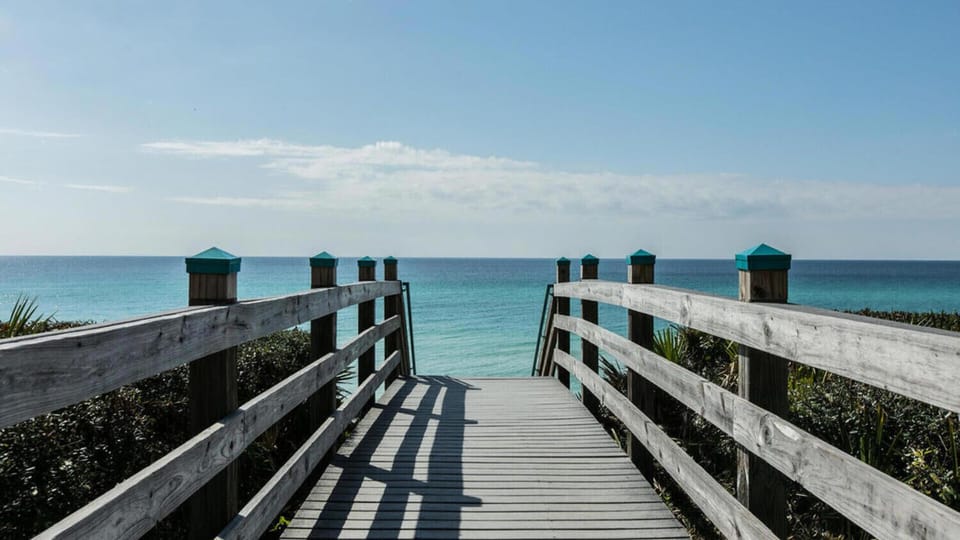 A wooden boardwalk leads through tropical vegetation to pristine turquoise waters and white sand beach.