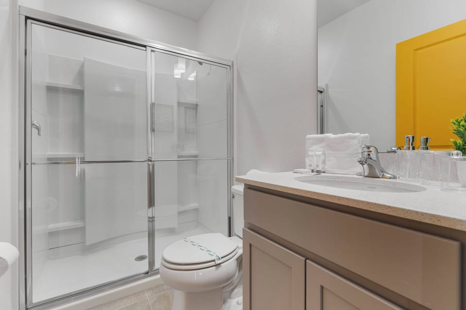 Experience a bright, functional bathroom with a modern vanity and ample counter space, complemented by neatly stacked towels. The inviting glass shower enclosure provides a refreshing retreat, accented by a cheerful yellow door.