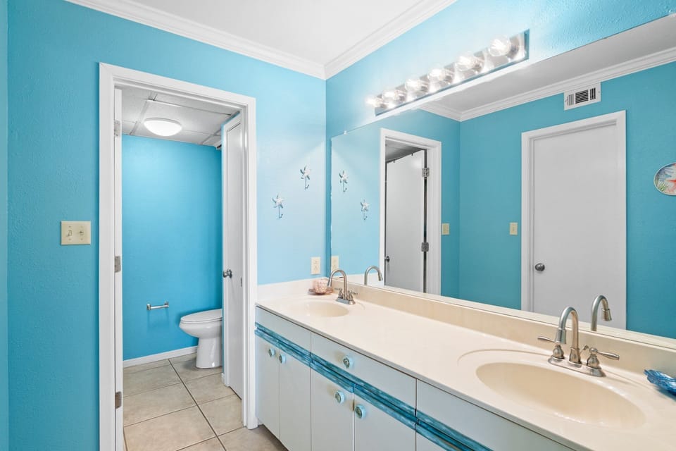 Spacious bathroom with double sinks and a separate toilet area in a bright blue setting.