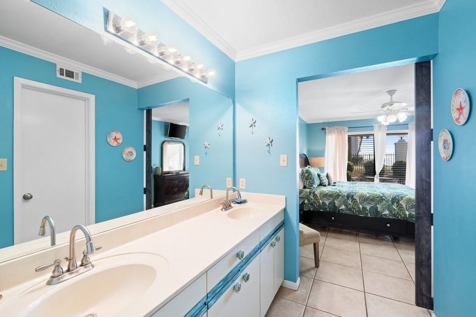 Bright bathroom with double sinks and a view into the bedroom featuring cityscape windows.