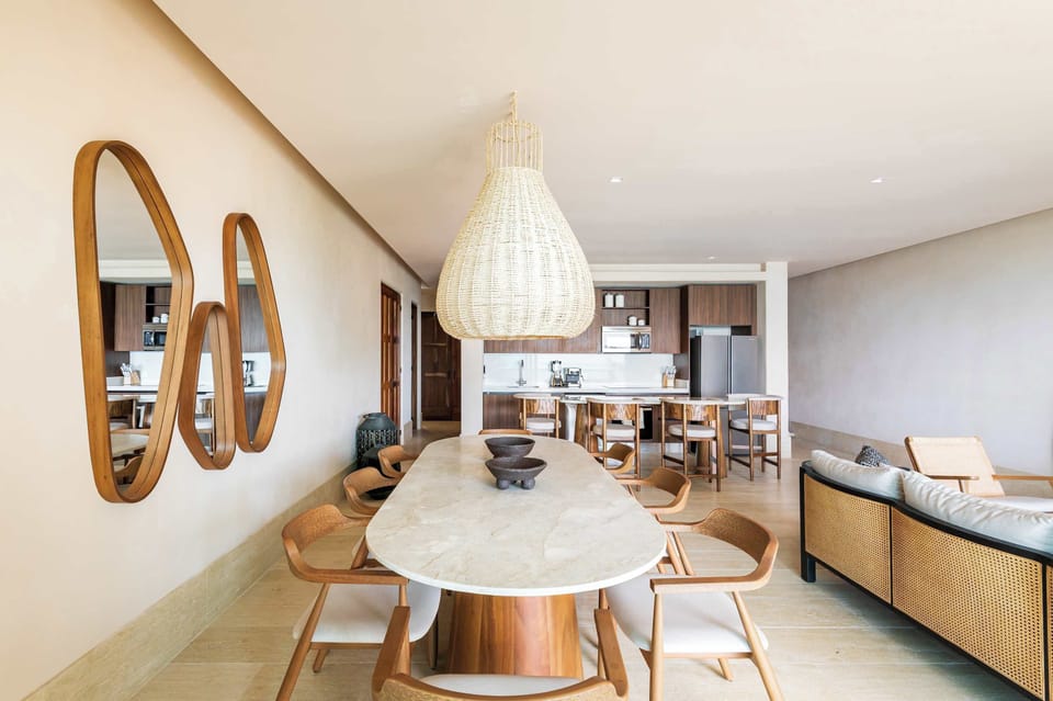 A bright dining area with an oval table, wooden chairs, and a woven pendant light, adjacent to a modern kitchen.
