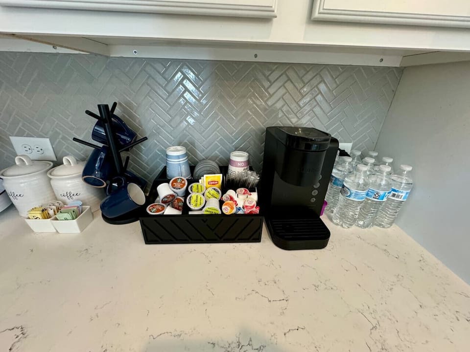 Coffee station with a black coffee maker, mugs, K-cups, creamers, sugar packets, and bottled water.