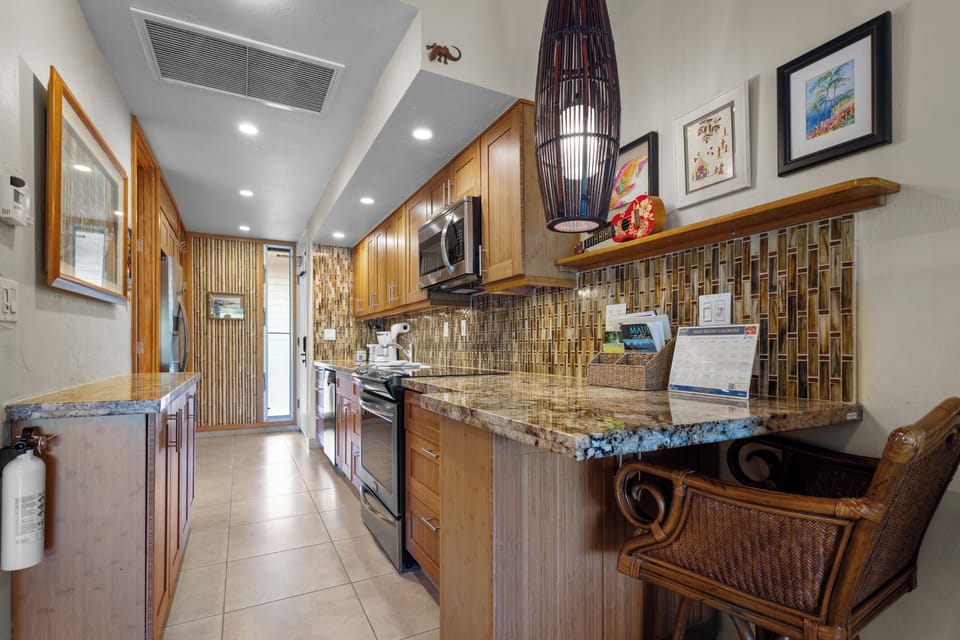 Step into your fully-equipped kitchen featuring warm wood cabinetry, granite countertops, and modern appliances. A woven bar seat invites morning coffee while tropical decor and stylish lighting create an island-inspired ambiance perfect for preparing meal