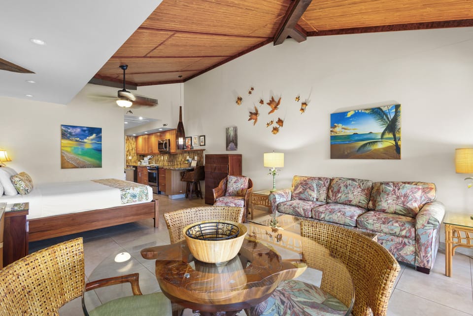 Relax in your open-concept studio with vaulted wood ceilings, a cozy living area, and tropical artwork creating a peaceful island vibe. The kitchenette and comfortable furnishings make it feel like home.