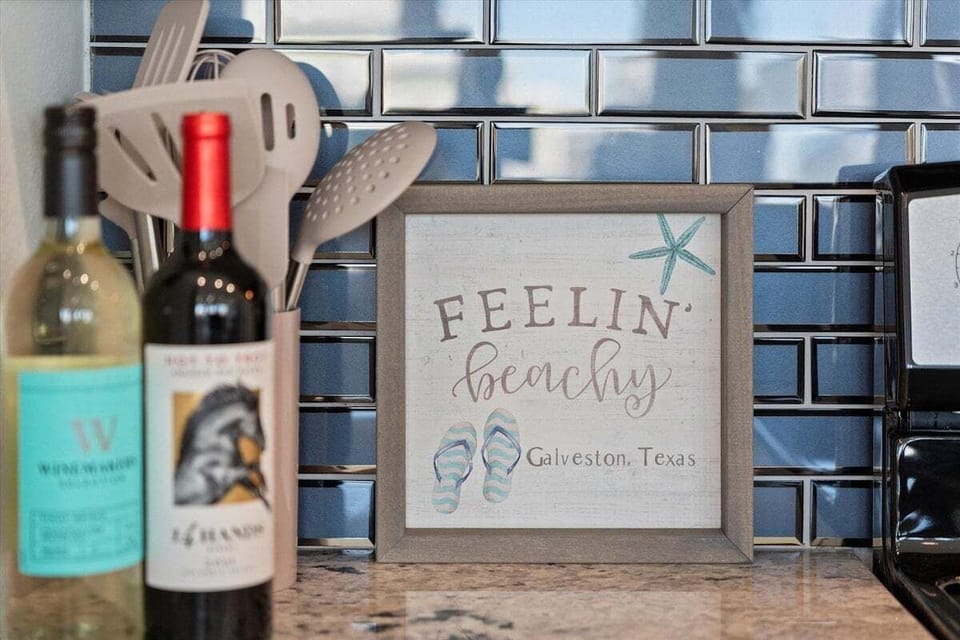 Coastal beverage display with wine bottles and decorative Galveston-themed accents set against a modern tile backsplash.