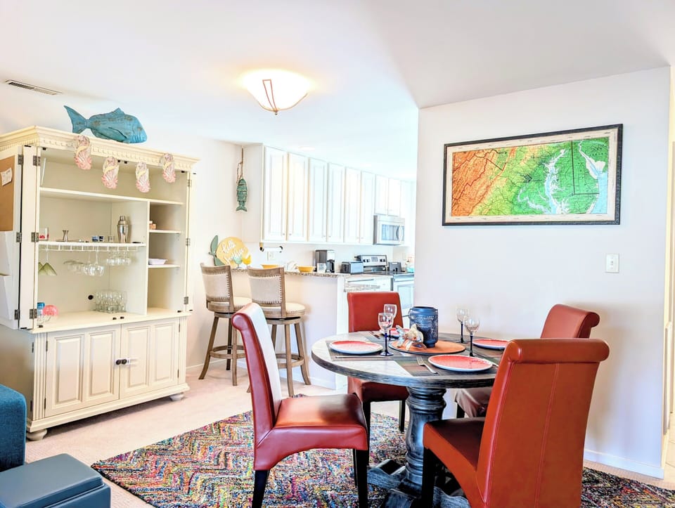 719A Treetop Lane - Dining and Hutch Perspective - Coastal dining space showcasing a round wooden table with four red leather chairs, a white storage hutch, and colorful abstract wall art near the breakfast bar area.