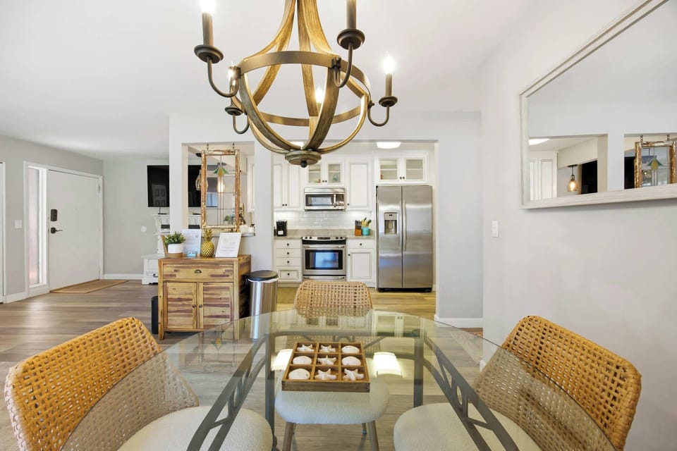 Wicker chairs and a glass-topped table sit beneath a curved wooden chandelier, creating a bright spot for morning coffee. Smooth stainless steel and white cabinetry gleam in the open kitchen, offering a modern space for shared meals.