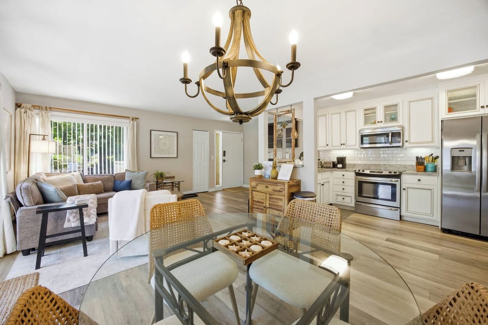 A glass dining table and rattan chairs anchor this airy open floor plan, where white cabinetry meets warm wood accents. Golden light spills across the wide-plank floors, connecting the modern kitchen to a cozy lounge perfect for long conversations.