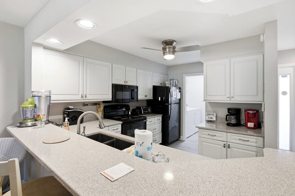 Speckled stone counters sprawl across this bright kitchen, offering a wide perch for morning coffee. Smooth white cabinetry and soft overhead beams create a clean, airy space for preparing shared meals.
