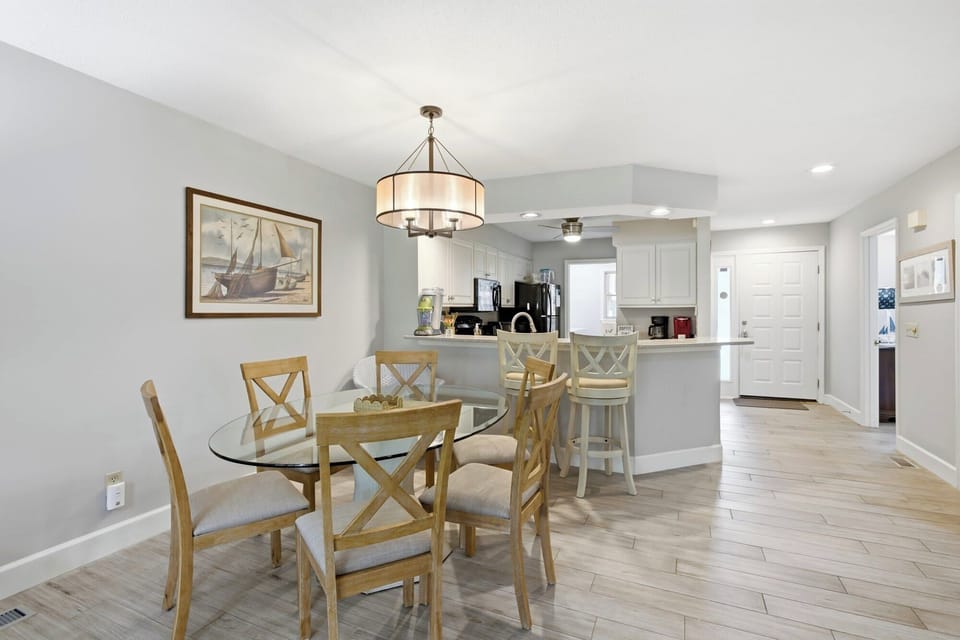 Oak chairs surround a glass table beneath a drum pendant light, creating a bright spot for shared meals. Pale wood floors and soft gray walls offer a calm, breezy backdrop that connects the dining area to a modern kitchen.