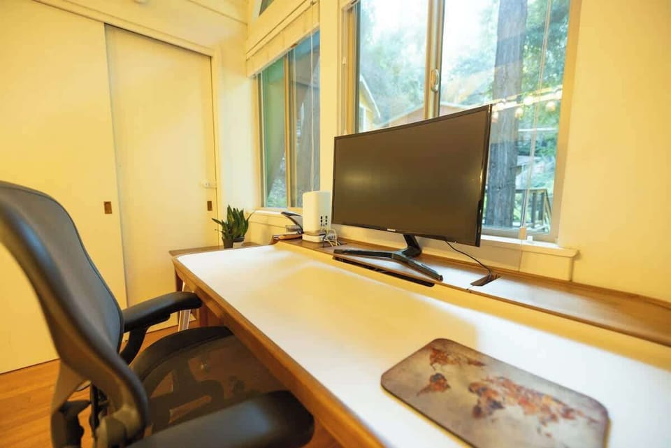 A spacious desk with large monitor and comfortable seating sits beside bright windows, offering a quiet and well-equipped workspace with calming outdoor views
