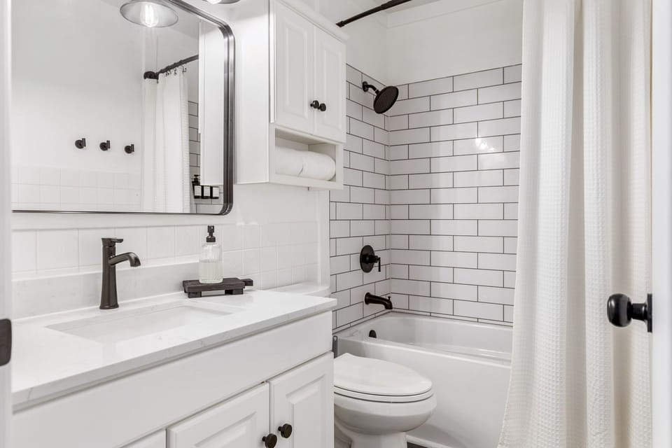 White bathroom with vanity, toilet, and bathtub-shower combination finished with subway tile and black fixtures.