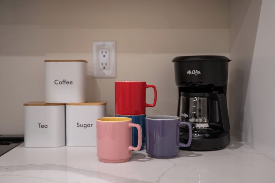 - Your morning starts right with a drip coffee maker, labeled containers, and cute ceramic mugs
- Rainbow toned cups bring cheerful color to the minimalist white counter
- All your caffeine essentials are thoughtfully organized and ready to use