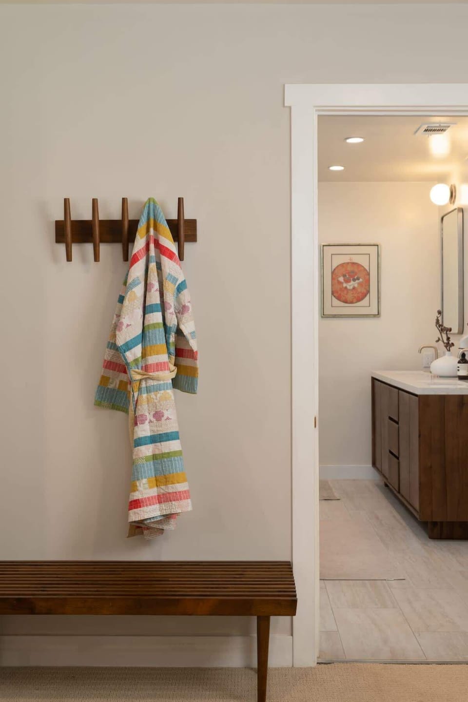 A charming hallway nook with a wooden bench and wall hooks offers a convenient spot for coats and towels, leading seamlessly into the bright and modern bathroom beyond