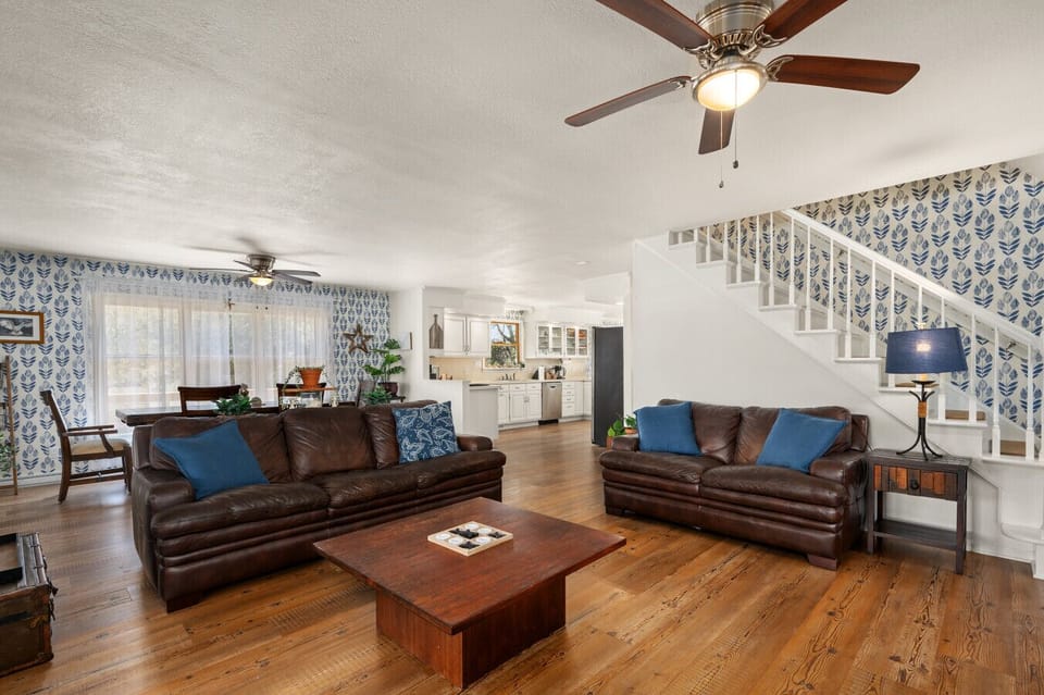 Bright living room w/ comfy sofas, ceiling fan, & open staircase, perfect for relaxing after a day out.