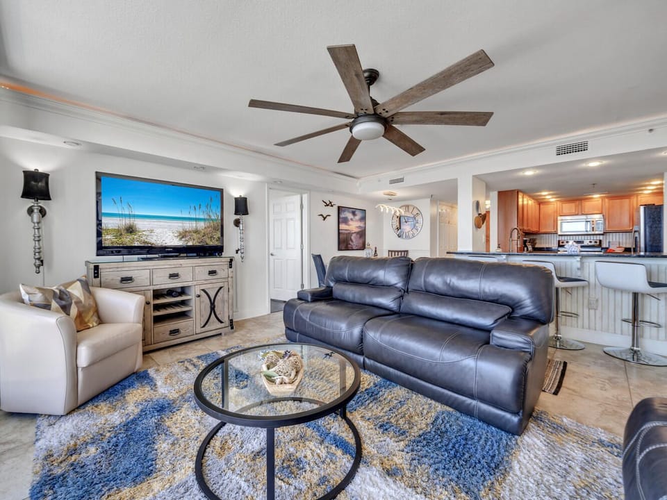 Relax in your spacious living room featuring leather seating and beach views on the large TV. The open kitchen awaits your culinary adventures.