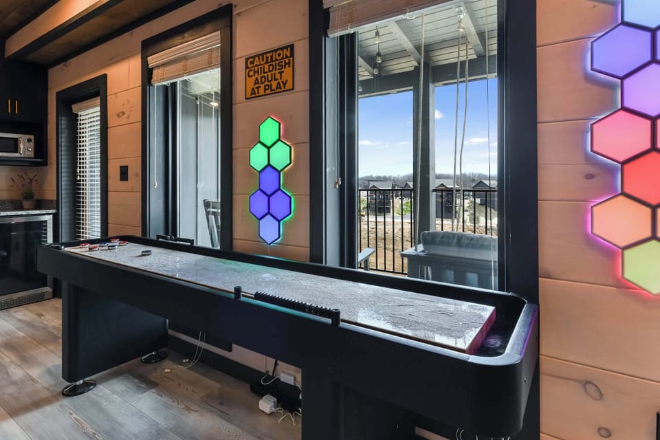 Game room with shuffleboard table and colorful hexagonal wall lights overlooking a balcony.