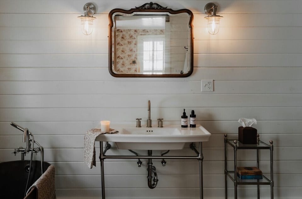 Rustic Yet Luxurious Bathroom Vanity – A classic farmhouse sink with elegant brass fixtures is complemented by warm lighting, an antique mirror, and thoughtfully curated toiletries.