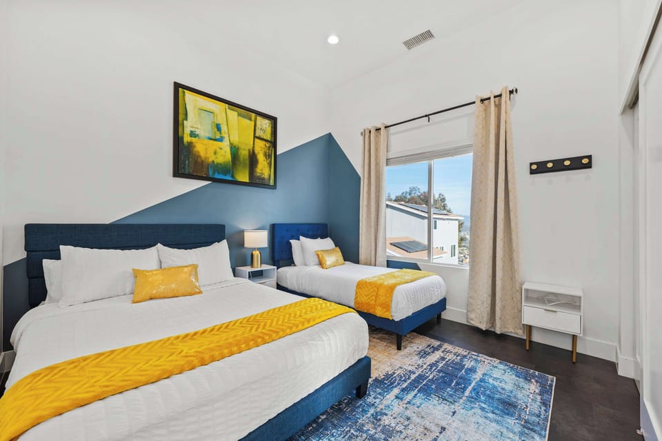 This bedroom showcases a bold blue and yellow theme with a queen and twin bed. A geometric accent wall, modern décor, and natural light create a cozy, contemporary, and inviting sleeping space.