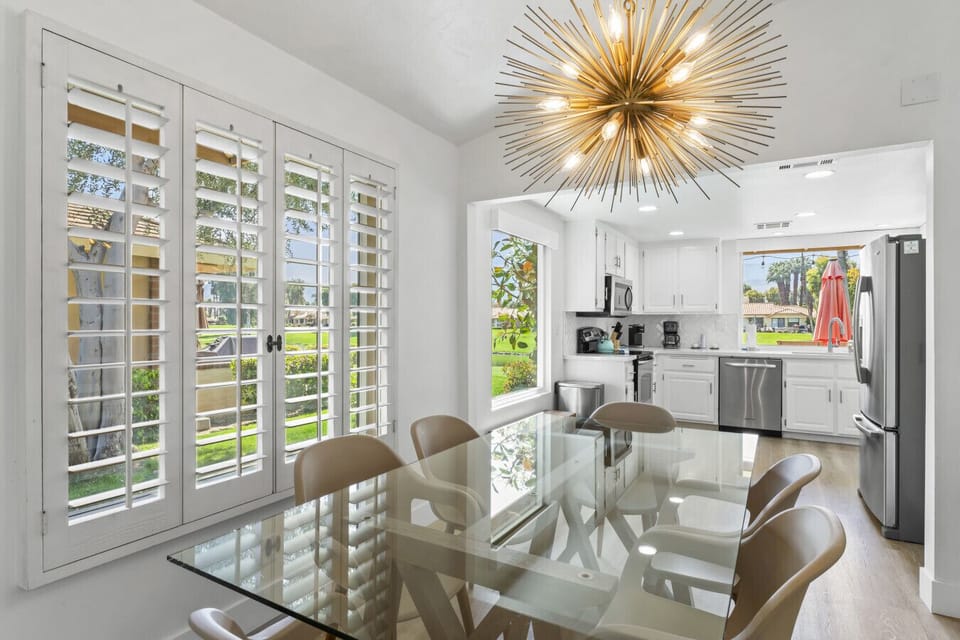 Elegant dining space w/ glass table, modern chandelier & shutters letting in soft natural light