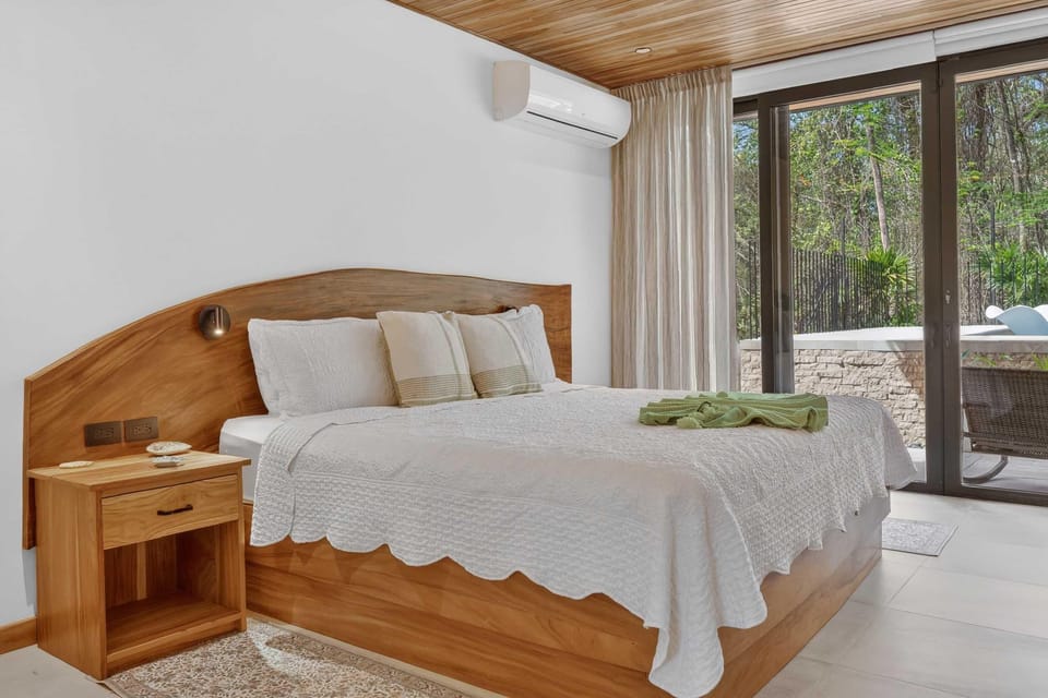 Natural wood headboard curves behind a king bed dressed in white quilted bedding, near glass doors to a patio.
