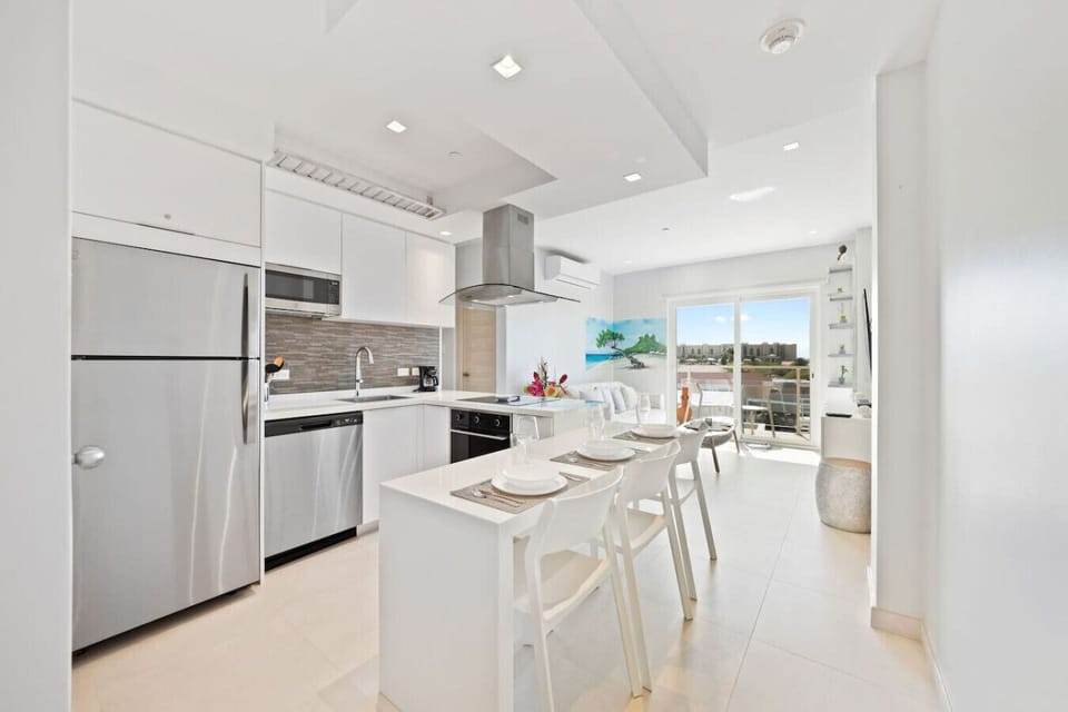 Modern kitchen with white cabinets, stainless steel appliances, and a gray tiled backsplash. The kitchen island doubles as a dining table with four white chairs. The space opens up to a bright living area and a balcony.