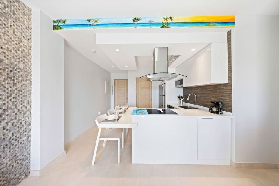 A modern kitchen and dining area with a clean, minimalist design. The kitchen features white cabinets and stainless steel appliances. A beach-themed mural is painted on a bulkhead above the counter.