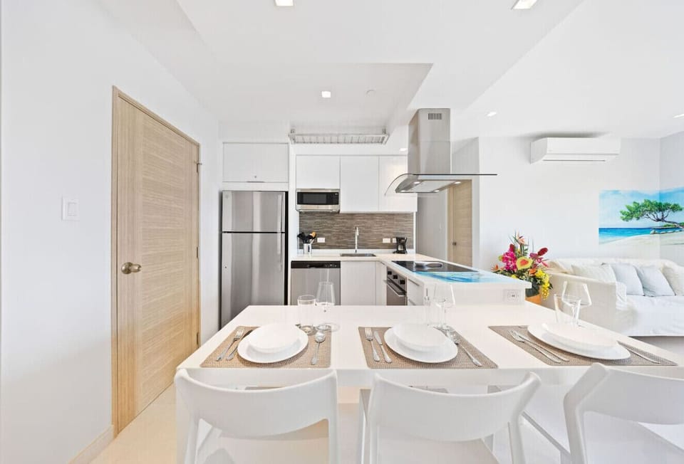 Modern kitchen and dining area with a white table set for four. The kitchen features white cabinets, stainless steel appliances, and a gray tiled backsplash. A large artwork of a beach scene is visible in the background.
