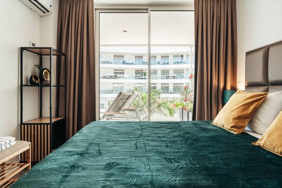 Wake up to sunshine and style—this elegant bedroom opens onto a private balcony, with rich green bedding, golden pillows, and sleek decor for the perfect blend of comfort and charm.