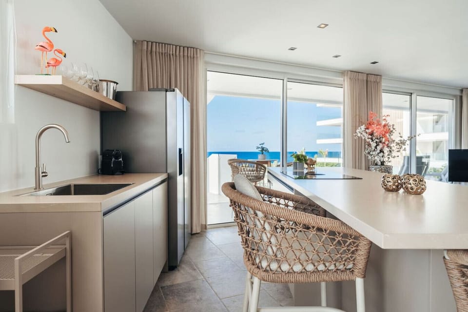 Coastal charm meets modern flair—this breezy kitchen features woven barstools, sleek surfaces, and ocean views that turn everyday dining into a scenic, sunlit experience.