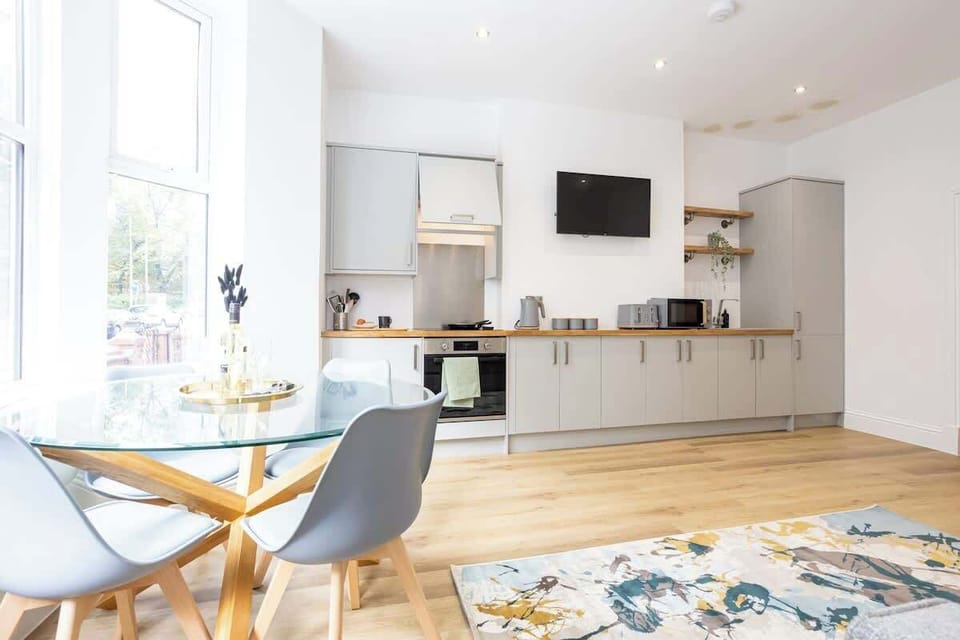 Bright open-plan kitchen and dining area featuring sleek grey cabinets, wooden countertops, and modern appliances. A round glass table with stylish chairs sits by large windows, while a colorful rug adds warmth and charm to this inviting space.