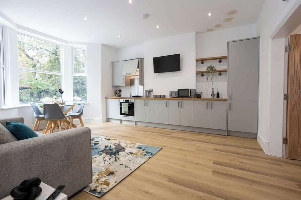 Bright open-plan kitchen and dining area featuring sleek grey cabinets, wooden countertops, and modern appliances. A round glass table with stylish chairs sits by large windows, while a colorful rug adds warmth and charm to this inviting space.