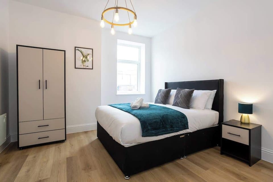 The room features a comfortable king bed with crisp white bedding and plush pillows, a tall wardrobe with dual-tone doors, and a bedside table. There’s a modern pendant light overhead, wooden flooring, and an ensuite bathroom .