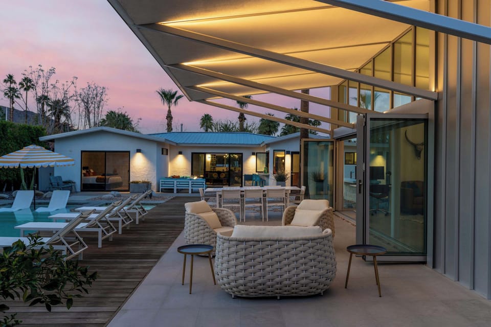 Twilight patio with lit retractable awning, woven lounge chairs, outdoor dining, and pool — the whole compound glowing at golden hour