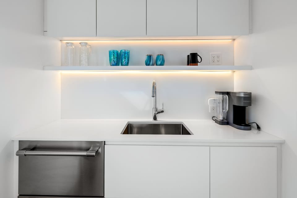 Suite kitchenette with white quartz counters, undermount sink, under-counter fridge, Keurig, and LED-lit open shelf — first coffee sorted