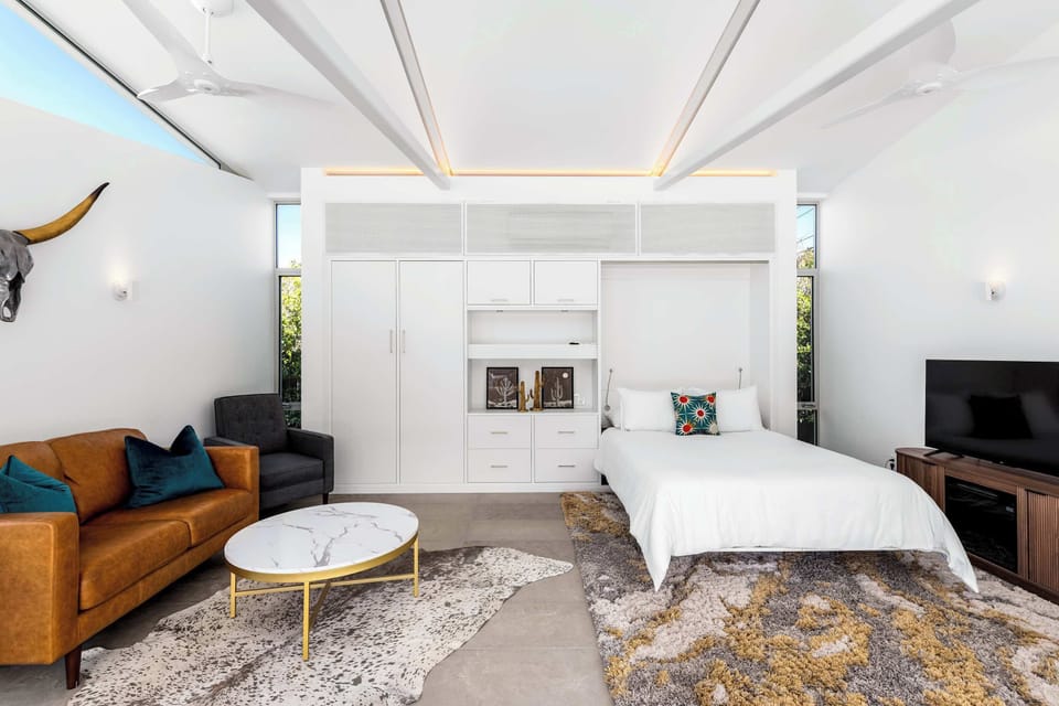 Bright guest suite with built-in wardrobe wall, platform bed, leather sofa seating area, and warm LED cove lighting — a polished private retreat