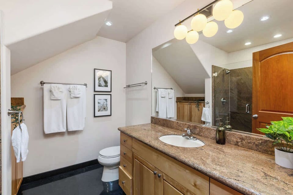 This bathroom blends functionality with elegance, featuring warm wooden cabinetry, a spacious granite countertop, and stylish lighting. The glass-enclosed shower adds a touch of luxury, while the neatly arranged towels. 