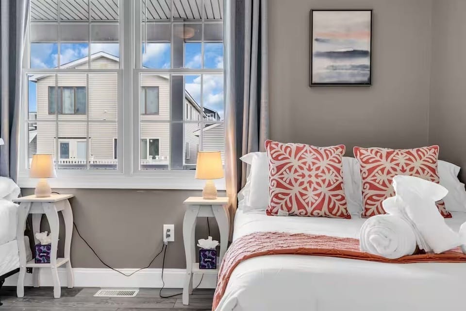 Bedroom with queen bed, red accent pillows, lamps, neutral linens, and natural window light.