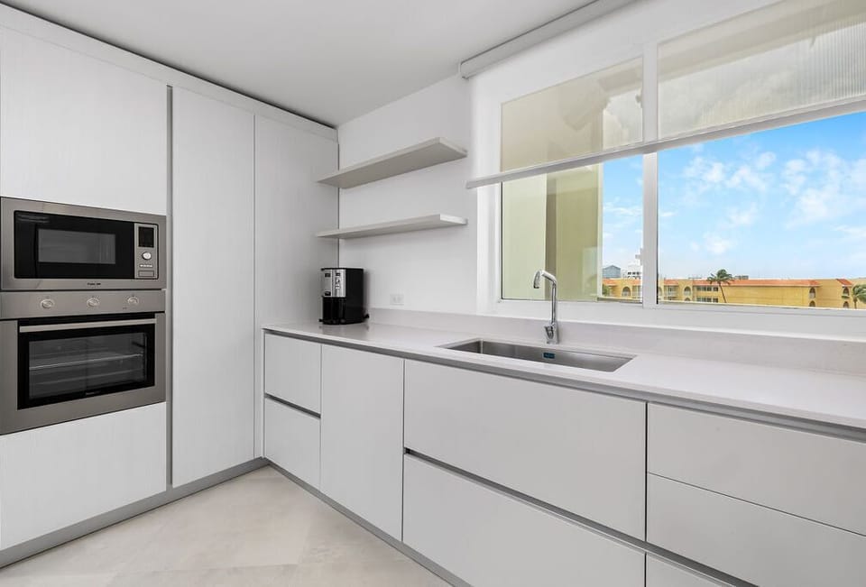 Your modern kitchen features sleek white cabinetry, stainless steel appliances, and a sunny window overlooking nearby buildings—perfectly outfitted for preparing delicious meals during your stay.