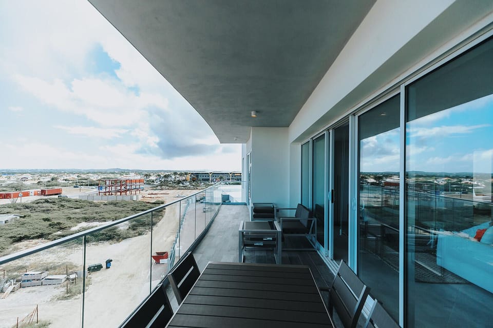 A covered balcony with contemporary outdoor seating overlooks a coastal landscape, offering views of the developing shoreline and expansive sky—a relaxing vantage point on arrival day.