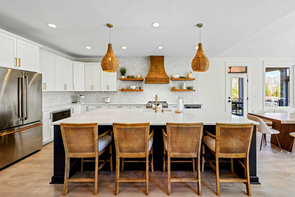 Chef's Kitchen - A massive quartz island seats four beneath woven pendants, with a statement wood hood and stainless appliances open to the dining and lake views.