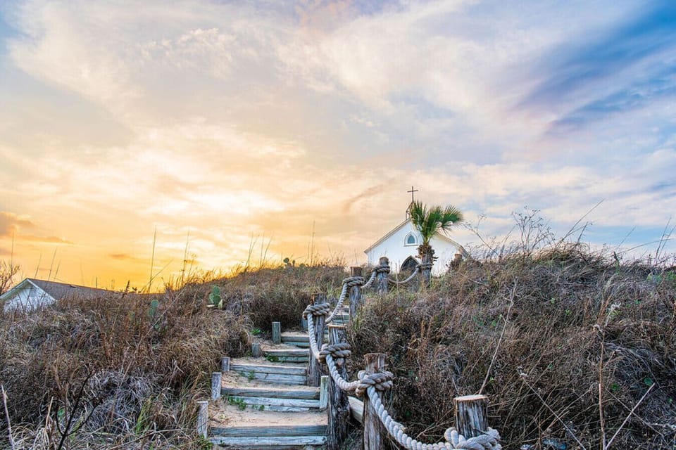 A small white chapel crowns coastal dunes, reached by weathered steps, glowing at sunset.