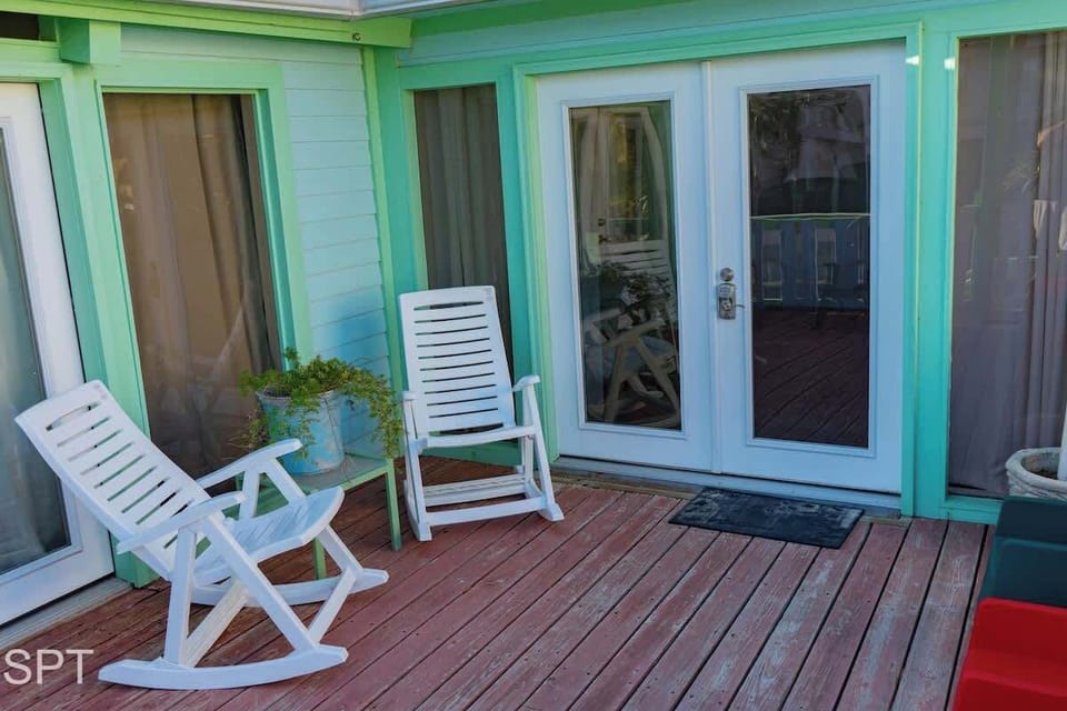 Cozy coastal porch featuring warm wood decking, classic white rocking chairs, and inviting double glass doors framed in soft mint. A small potted plant and relaxed outdoor setup create a charming spot to unwind and enjoy the breeze.
