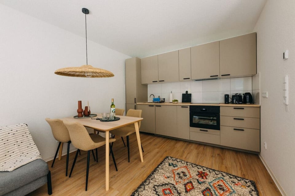 Moderne Küche mit warmen Tönen und kleinen Details. Perfekt ausgestattet mit Backofen, Spüle und Kaffeemaschine – hier kannst du dich wie zu Hause fühlen! Der gemütliche Essbereich lädt zum gemeinsamen Genießen ein. Willkommen in deinem Rückzugsort!