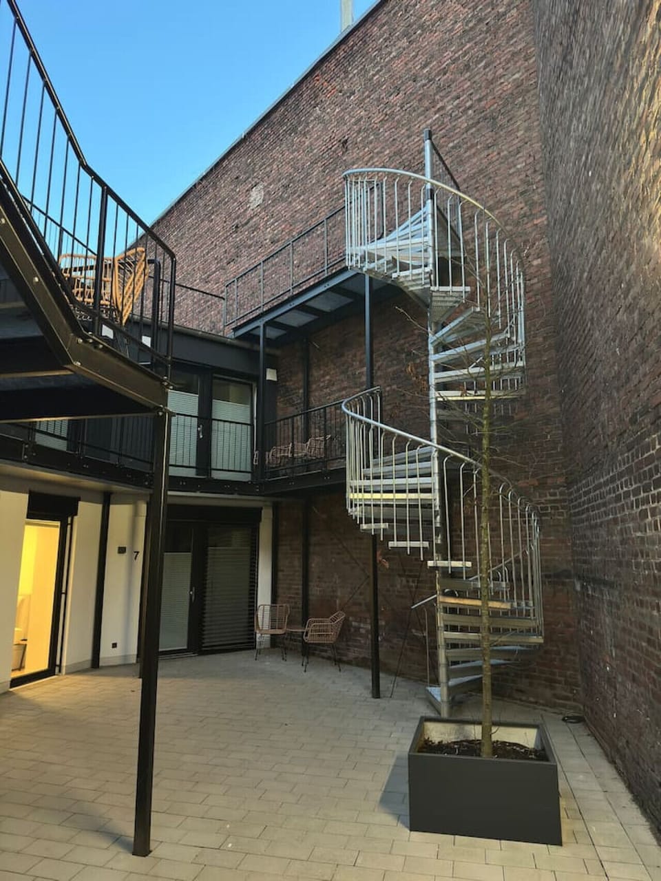 Urbaner Charme trifft auf modernes Design: Der begrünte Innenhof mit stilvollen Sitzmöglichkeiten und einer markanten Wendeltreppe verbindet Altbauflair mit zeitgemäßem Wohngefühl – ein perfekter Rückzugsort mitten in der Stadt.