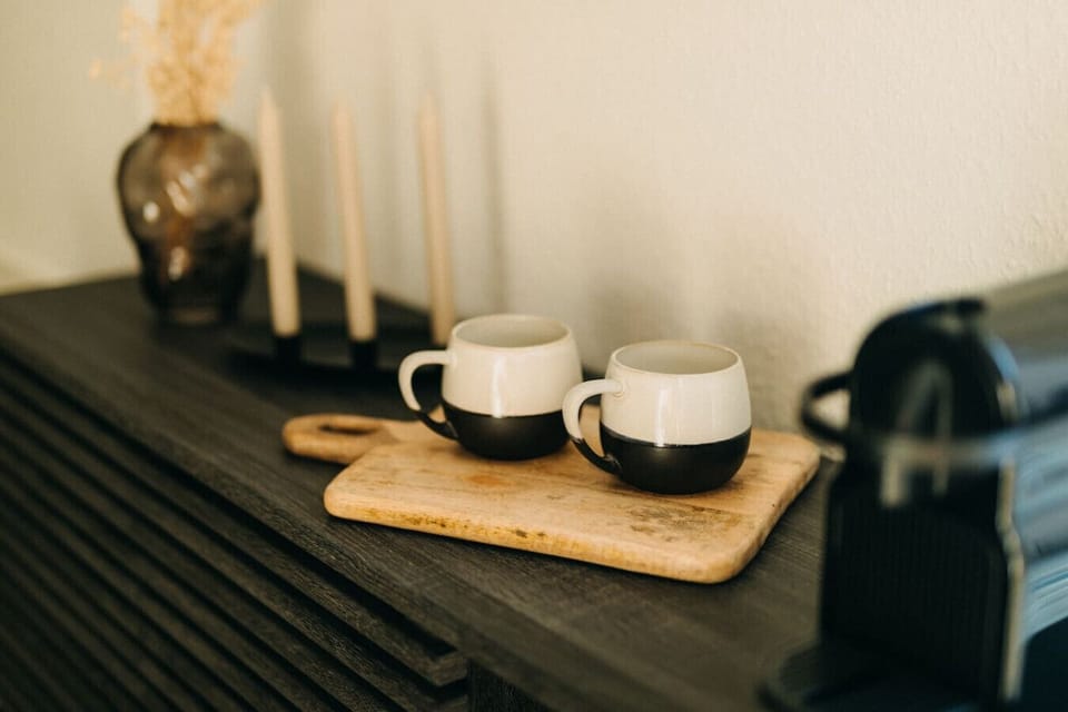 Morgenritual mit Stil: Zwei stilvolle Keramikbecher warten auf Ihren ersten Espresso des Tages. Die harmonische Kombination aus Holz, Keramik und dunklem Interieur sorgt für eine gemütliche Atmosphäre – perfekt für einen entspannten Start in den Tag.