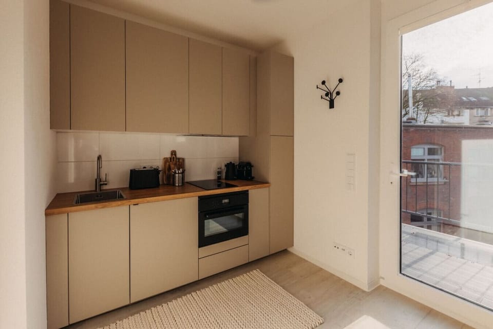 Die moderne Küche in warmen Erdtönen kombiniert stilvolle Schränke mit einer hochwertigen Holzarbeitsplatte. Voll ausgestattet mit Backofen, Kochfeld und Kaffeemaschine, lädt sie zum Kochen ein. Die großen Fenster sorgen für viel Tageslicht.