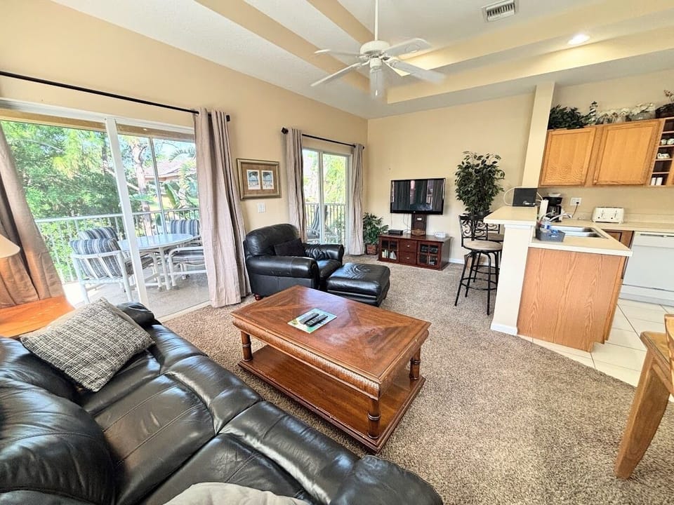 Comfortable living area with high ceilings, tile flooring, cozy seating, and two sets of sliding glass doors that bring in natural light and lead out to the private screened balcony.