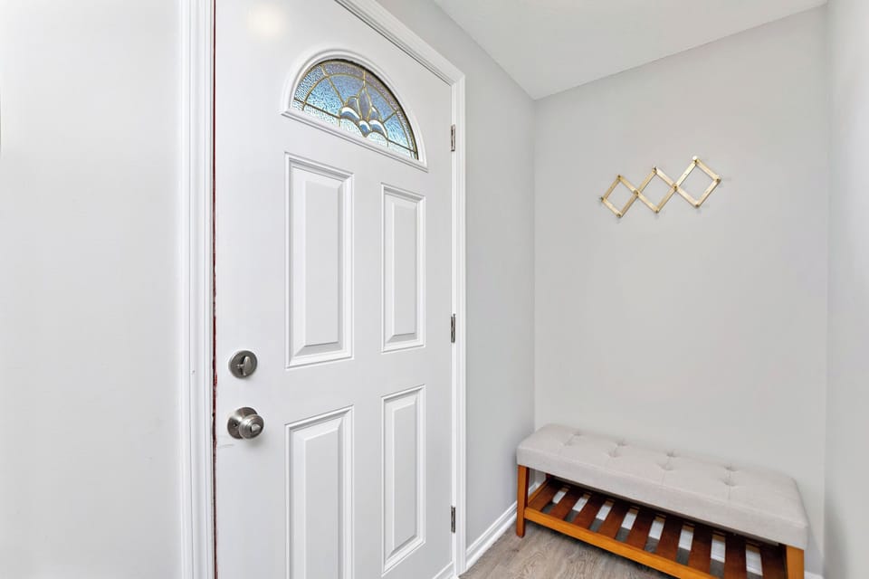 Inviting entryway with a white door featuring a stained-glass window. A wooden bench with a cushioned seat provides a place to sit and remove shoes. A coat rack hangs on the wall, perfect for storing coats and hats. 