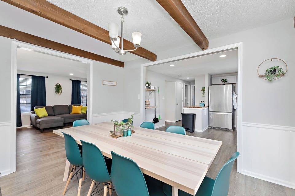 Spacious dining room with a large wooden table and teal chairs. Features a chandelier, wall decor, and an open-concept view of the living room and kitchen. Perfect for family meals and entertaining guests.
